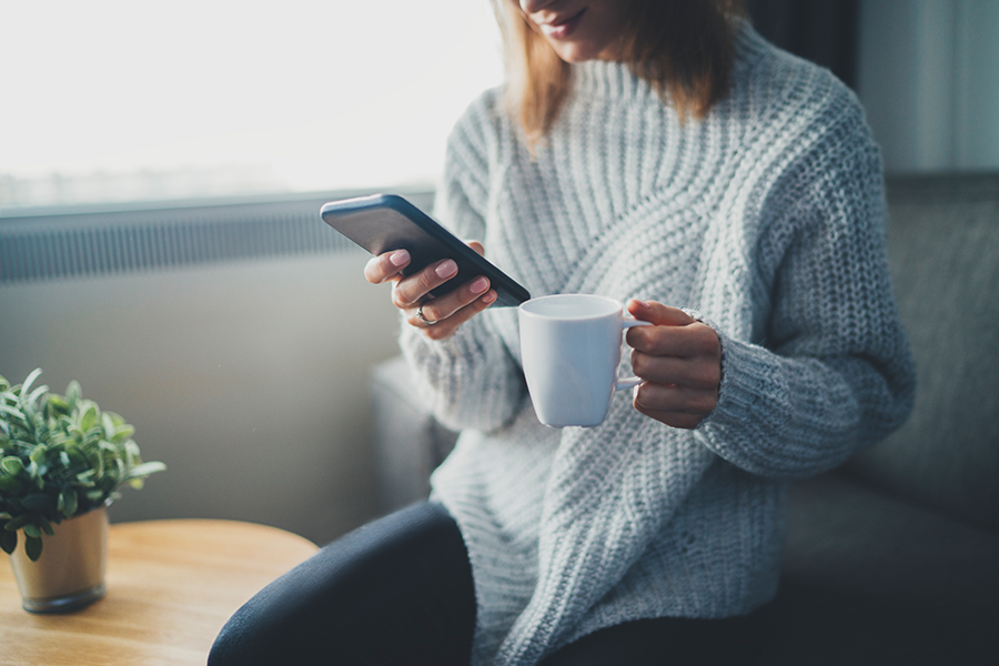 스웨터를 입은 여성이 소파에 앉아 커피를 마시며 스마트폰을 보는 사진
